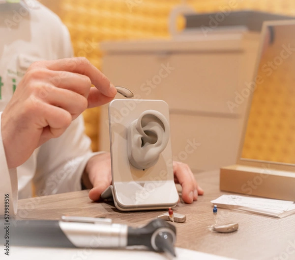 Fototapeta Doctor in his audiology office holding a hearing aid and in the other hand a model of a silicone ear. On his table is another hearing aid and medical devices.