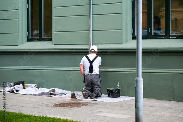 Fototapeta Maler und Lackierer arbeitet mit Pinsel und Farbe an einer Fassade und Regenrinne eines Hauses an einem Bürgersteig, viel Textfreiraum