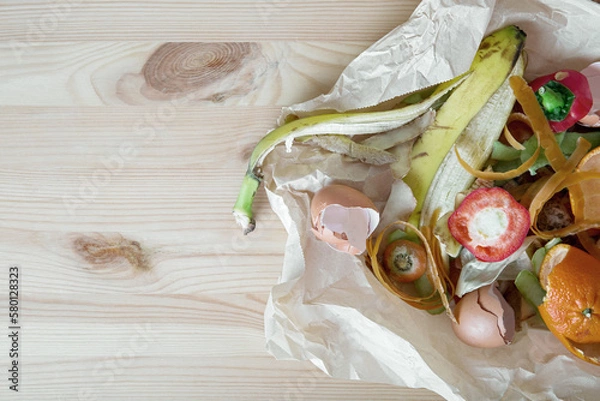 Fototapeta Compost from peelings of vegetables, fruits and egg shells on craft paper on a wooden table. The concept of waste-free consumption and eco-friendly lifestyle. View from above.