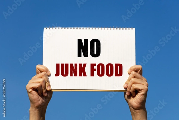 Fototapeta No junk food text on notebook paper held by 2 hands with isolated blue sky background. This message can be used as business concept to encourage audience not to eat junk food.