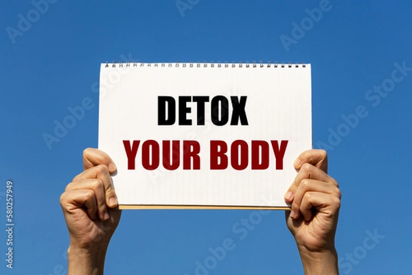 Fototapeta Detox your body text on notebook paper held by 2 hands with isolated blue sky background. This message can be used as business concept about detoxing your body.