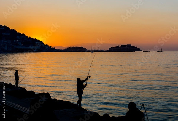 Fototapeta Silhouette of fisherman fishing at sunset in front of the sea. Beautiful and peaceful landscape of sunset on the coast with fishermen. Vacation in nature.