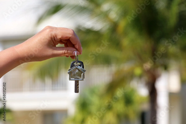 Fototapeta Real estate agent, home keys in female hand on background of house surrounded by palm trees. Buying or rental a villa on ocean coast, removal to tropical country