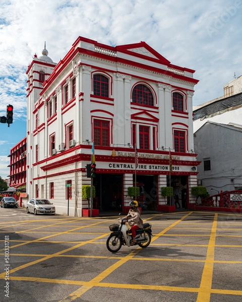 Fototapeta Colonial fire station building. In front of the building on the street a woman in a helmet on a motorcycle. sunny day. georgetown. Penang. Malaysia