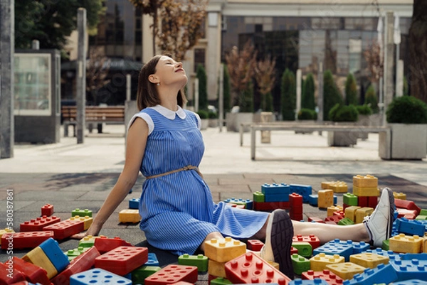 Fototapeta A beautiful pregnant woman in a blue dress playing with big colorful cubes on a children playground while sitting and watching the cubes. Pregnancy creative concept
