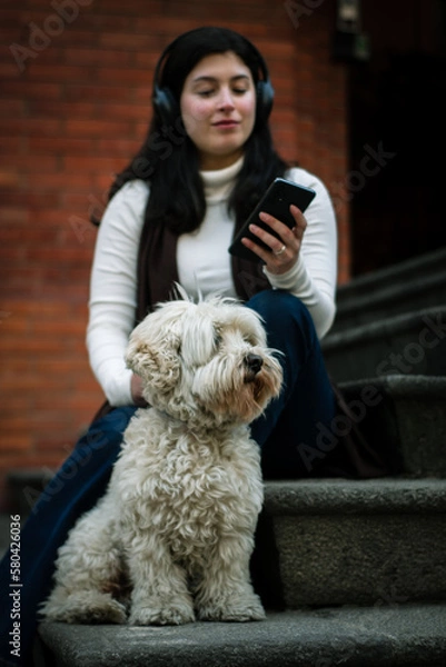 Fototapeta The fluffy white dog looks into the distance. The owner of him is back in the background listening to music. Relaxed girl with her dog listening to music from the cell phone.