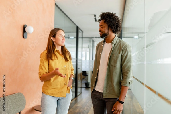 Fototapeta Young enthusiastic female colleague explains new strategy to male employee and gesturing. Indoor portrait of diverse work team discussing, walking through hallway in modern coworking office space