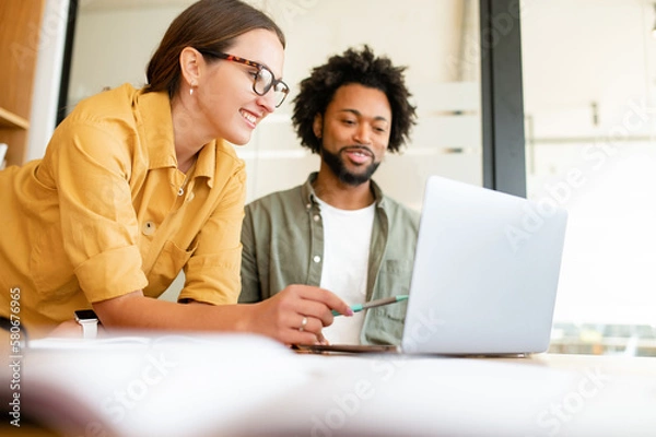 Obraz Two colleagues have pleasant conversation sitting side by side in modern open space office in front of laptop, smiling and enjoying friendly working atmosphere, brainstorming together, solving tasks