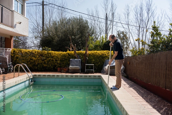 Fototapeta man cleaning swimming pool with manual vacuum cleaner, pool maintenance after winter to prepare the water for summer, private house in urbanization