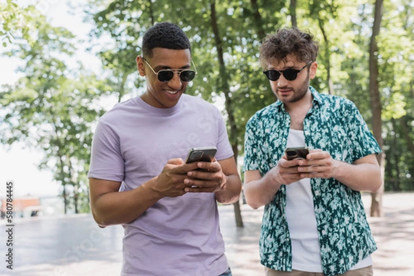 Fototapeta Smiling interracial friends in sunglasses using smartphones in park.