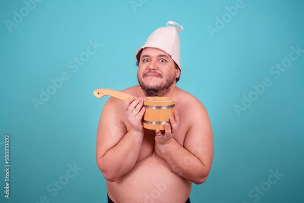 Obraz Funny fat man in a hat and with a sauna ladle. Blue background. Taking a shower.