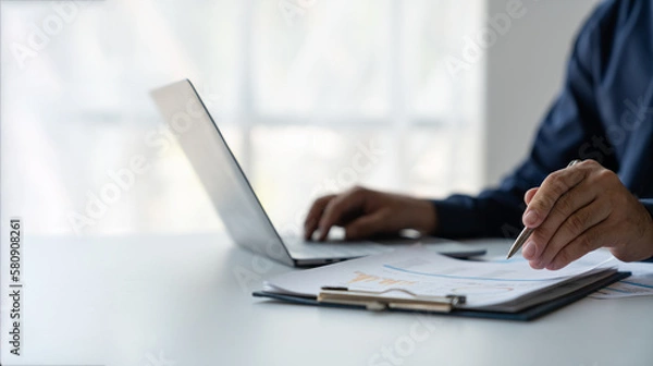 Obraz young business man Recording data on laptop, comparing details from graphs. Business income charts in the areas of real estate, tax, management, technology marketing in a private office.