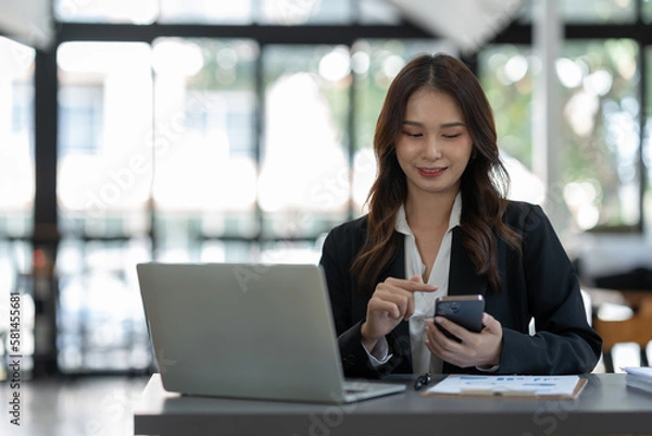 Fototapeta Attractive Asian businesswoman using a mobile phone at the office for making contact Talk about online business. and financial income happily.
