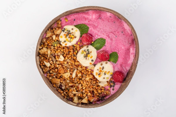 Fototapeta Smoothie bowl in a coconut plate with raspberries, yogurt, bananas, nuts, mint and roasted buckwheat. White background, top view.