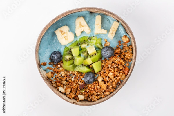 Fototapeta Summer blue smoothie bowl with yogurt, kiwi, blueberries, banana lettering Bali, nuts and buckwheat. A plate of coconut on a white background. Top view.