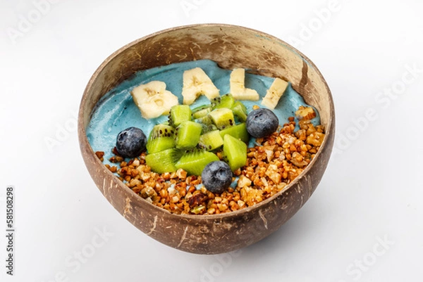 Fototapeta Summer blue smoothie bowl with yogurt, kiwi, blueberries, banana lettering Bali, nuts and buckwheat. A plate of coconut on a white background. Close up 3/4