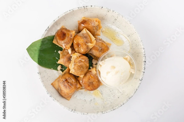Fototapeta Bananas in rice paper with cinnamon, Jerusalem artichoke syrup, peanuts and ice cream. White ceramic plate on a white background. Top view.