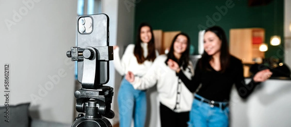 Fototapeta Close-up of a smartphone on a tripod filming 3 young white girls dancing, at home. Concept of making a living with videos on the Internet by being an influencer. Popularity and celebrity online