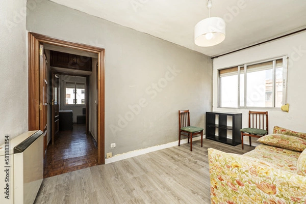 Fototapeta Living room furnished with a solitary sofa in a patterned fabric of dubious taste, sapele wooden doors and gray wooden floors, poorly painted walls and a ceiling lamp