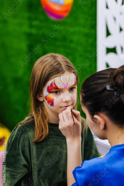 Fototapeta A professional make-up artist, artist paints with a brush on her face with multi-colored paints face painting, drawing, children's makeup for a little girl model. Photography, art concept, lifestyle.