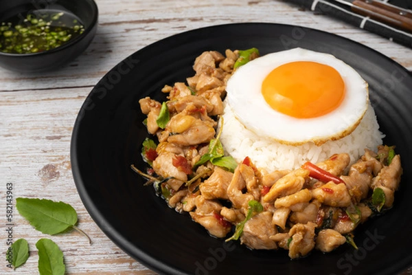 Fototapeta Closeup of stir fried basil and chicken on rice with fried egg in black dish on wooden table.