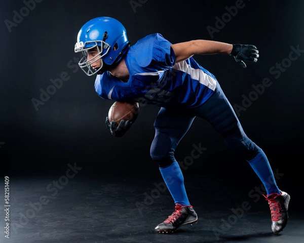 Fototapeta Full length portrait of a man in a blue american football uniform against a black background. Sportsman in a helmet with a ball. 