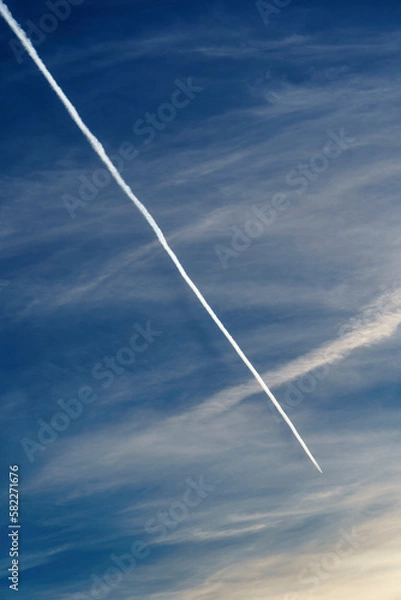 Obraz An airplane flying through a clear blue sky, leaving a long and persistent white contrail behind it. In the background, some soft and diffuse clouds add a touch of texture to the image. The bright blu