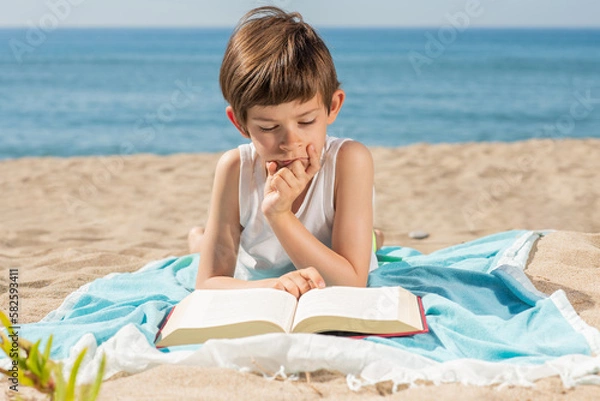 Fototapeta Child immersed in reading a book on the towel on the beach on a sunny day with the sea in the background