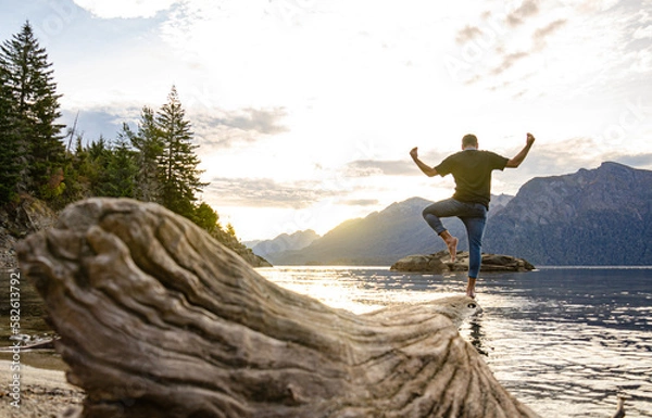 Obraz persona sobre un árbol al borde del agua de un lago en la Patagonia Argentina al atardecer con montañas de fondo mirando el paisaje