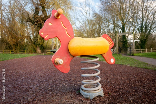 Fototapeta Colourful children's rocking horse on springs over wood chip floor in outdoor playground with trees.