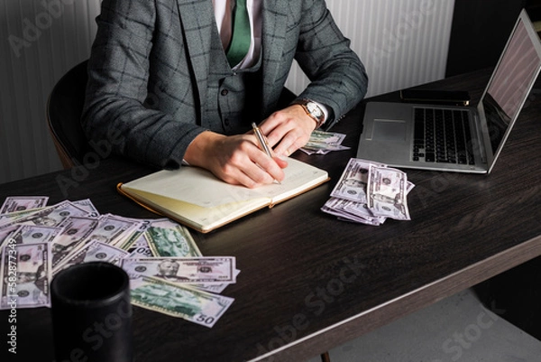 Fototapeta Close-up of a young serious man in a gray formal suit sits at his desk surrounded with cash and takes a note in his notebook. A businessman at work.