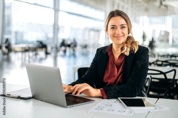 Obraz Successful confident caucasian business woman dressed in elegant wear, executive, recruitment, product manager, sits in a modern office, works in a laptop, looks at camera, smiling friendly
