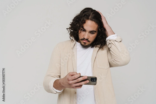Fototapeta Upset hispanic man playing online game on mobile phone isolated over white wall