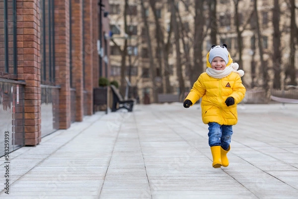 Fototapeta Portrait of a small child 4 years old in yellow rubber boots and a jacket. The kid walks in the park in the spring. Photo of spring and autumn holidays.
