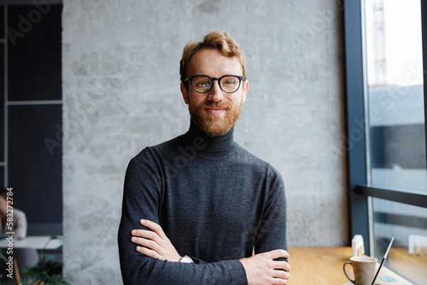 Obraz A young red-haired guy, a programmer or an entrepreneur in glasses, in a stylish cafe Looks at the camera and smiles. Freelancer works remotely. Online communication. small business.
