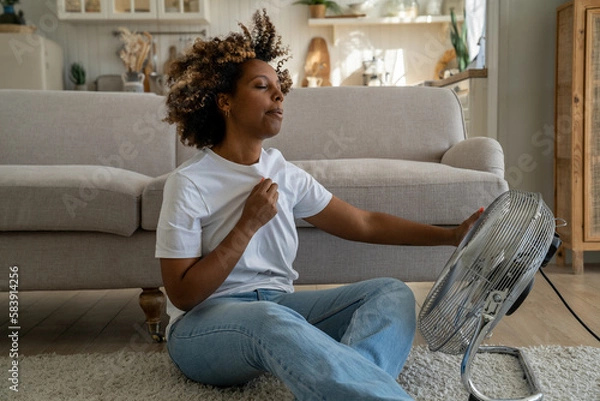 Fototapeta Summer heat. Young African American woman cooling down by ventilator at home, feeling unwell with high temperature during hot weather, sitting on floor in front of electric fan during extreme heatwave