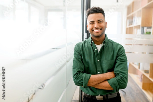 Fototapeta Handsome indian male office employee standing with his arms crossed, looking at the camera and laughs. Portrait of cheerful young man wearing smart casual shirt in modern coworking space