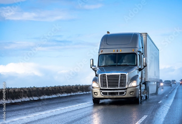 Obraz Gray big rig semi truck with turned on headlights transporting cargo in dry van semi trailer running on the wet evening road with rain and reflection on the road surface