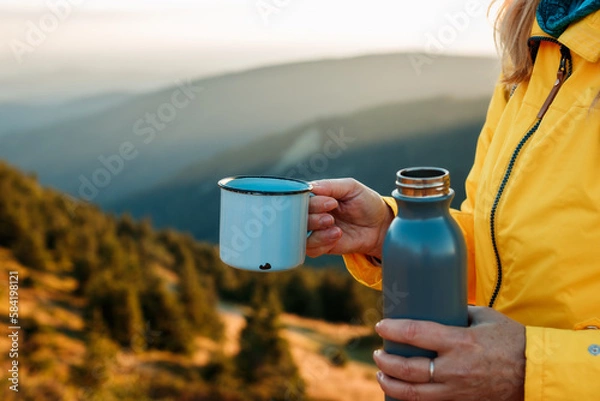 Fototapeta Refreshment after successfully climbed mountain summit. Female hiker drinking hot drink from travel mug and enjoying view at mountains during sunset
