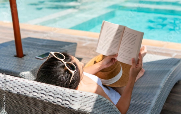 Fototapeta Happy girl lying down on sunbed while reading a book near Swimming Pool on travel holidays vacation, wellness Lifestyle concept. Young Asian woman relaxing at hotel on weekend. Copy space