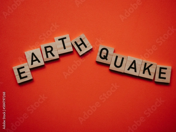 Fototapeta Inscription earthquake from vibration moves on background. Concept of earthquake, destruction and shifts of  earth's crust. Wooden square blocks with letters are laid out on  orange sheet. Slow motion