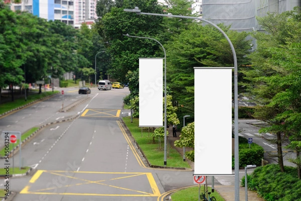Fototapeta Hanging posters by the road in the city; blank vertical advertising banners on street lampposts, against lush green trees and plants. For OOH out of home template mock up