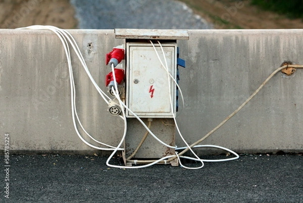 Fototapeta Heavy industrial outdoor metal electrical high voltage box with multiple outdoor electrical outlets and safety switches connected with thick electrical cables mounted on concrete wall near paved road 