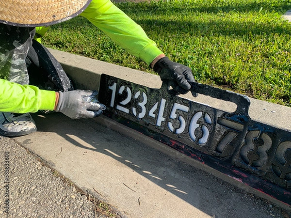 Obraz City worker painting address on street curbs using stencil numbers with white and blank paint
