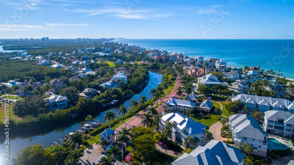 Fototapeta Aerial Perspective from Drone Featuring the Barefoot Beach in Bonita Springs, Florida. Blue Water Coastline with Clear Sky and Wispy Clouds in the Background and Clusters of Homes in the Foreground