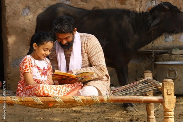 Obraz Happy rural Indian father and daughter sitting together on a cot. Father is teaching his daughter how to read with a book in his hand