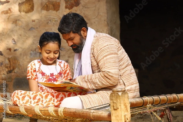 Obraz Happy rural Indian father and daughter sitting together on a cot outside their cottage/home/house. Father is teaching his daughter how to read with a book in his hand