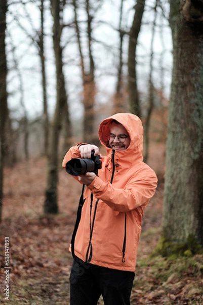 Fototapeta A young and smiling male while taking photos of the forest around him. Progressive photographer with a modern and expensive mirrorless camera. A man holding a camera with a telezoom lens in his hands.
