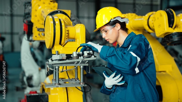Obraz Caucasian mechatronics engineer is inspecting a lot of robotic arms in warehouse before being used in factory. A female industrial worker is using a tablet to record the results of examining machines.