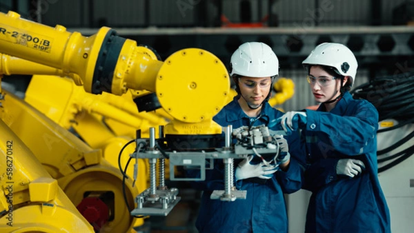 Obraz Two Caucasian mechatronics engineers are assisting in inspecting a lot of robotic arms in the warehouse. Female industrial workers are recording the results of examining machines on a tablet together.
